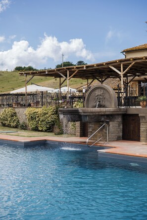 Une piscine agrémentée d’une fontaine en pierre, entourée d’un patio couvert avec des sièges d’extérieur.