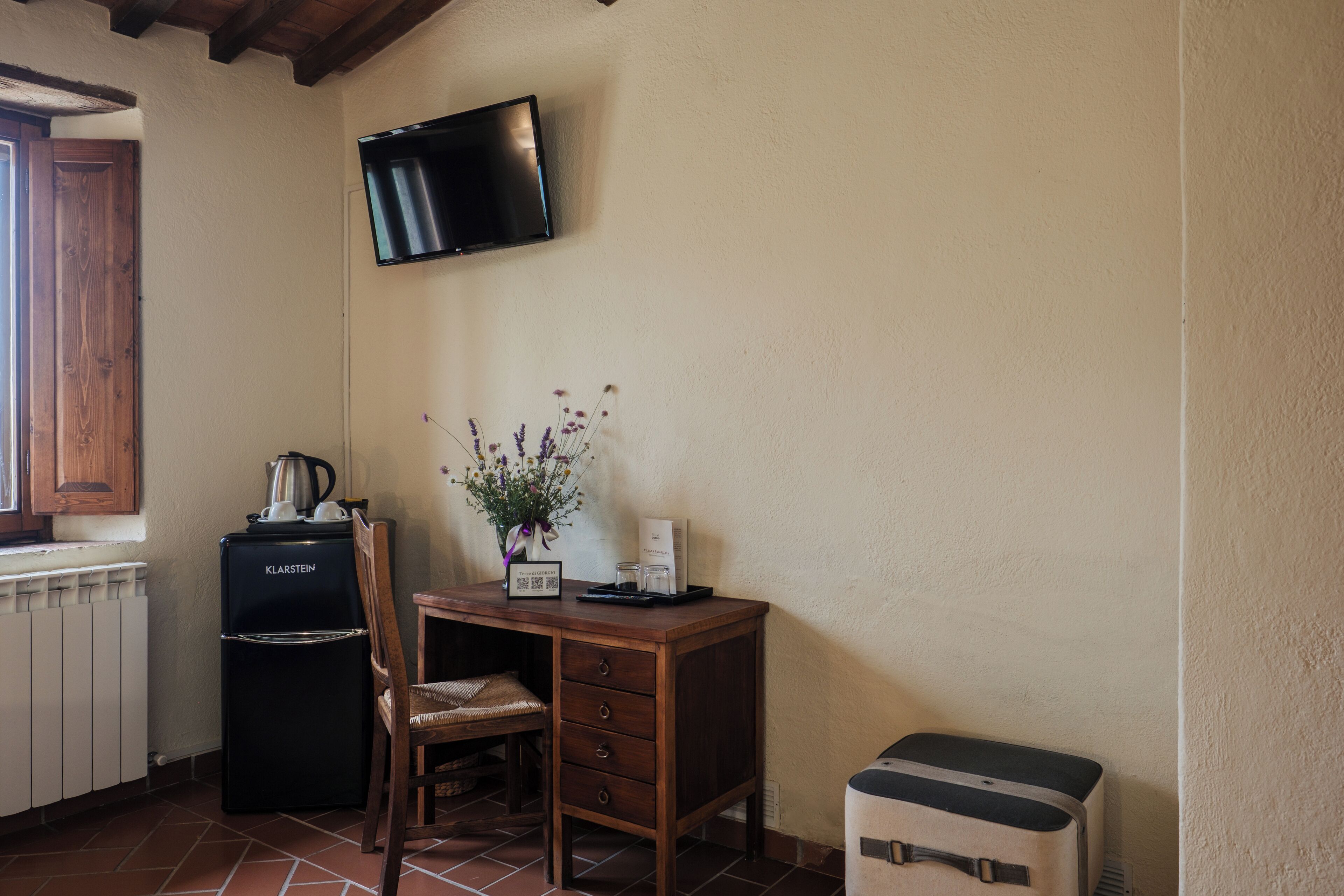A small kitchenette with a black refrigerator, a wooden chair, a wooden desk, a television mounted on the wall, and a small table with a vase of flowers.