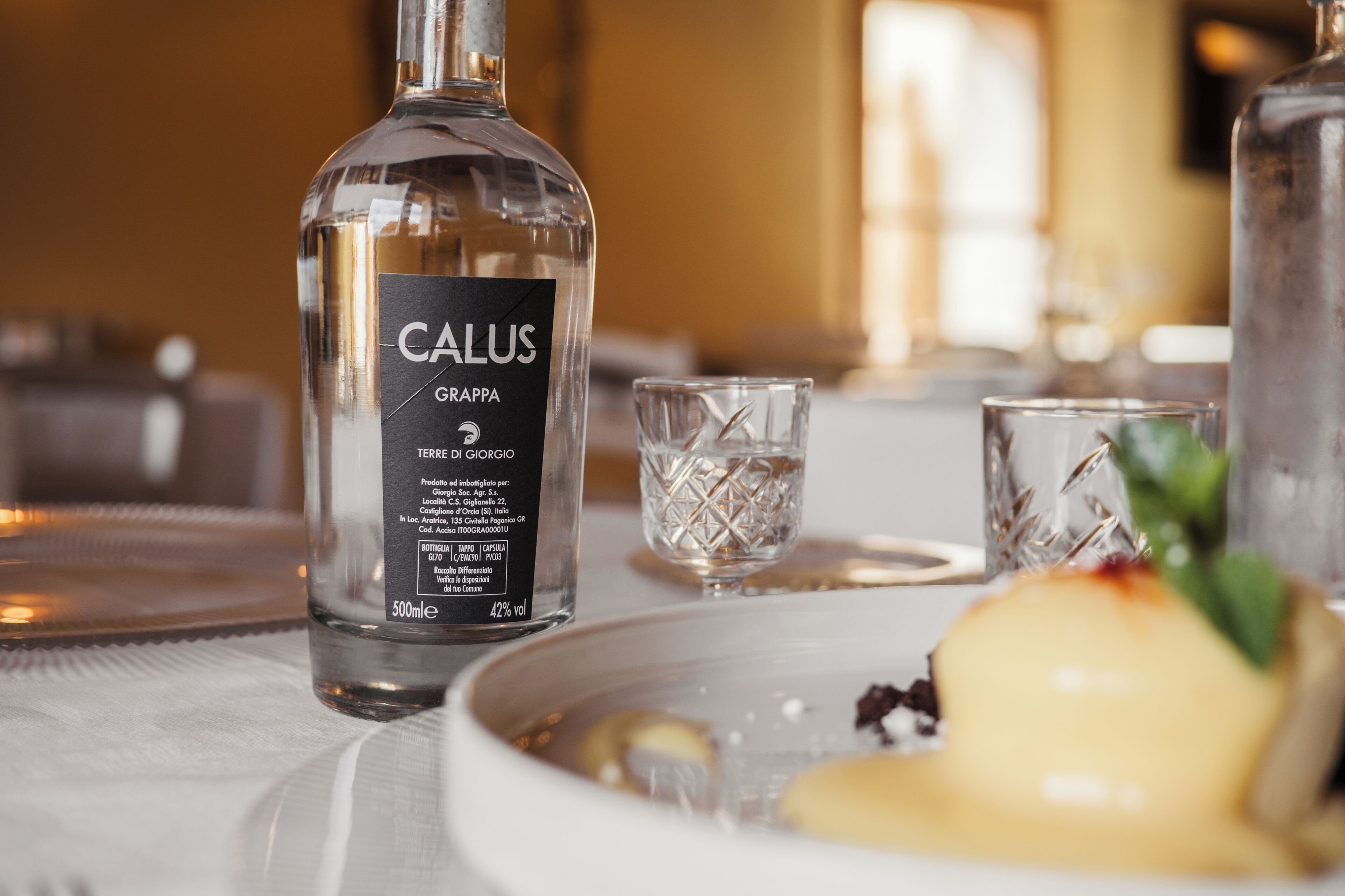 A bottle of CALUS Grappa on a table with a plate of food and two glasses.