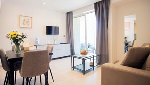 A modern living room with a dining area, a flat-screen TV, and a balcony with curtains. - Bentley Holiday Apartments - West One (Gibraltar)