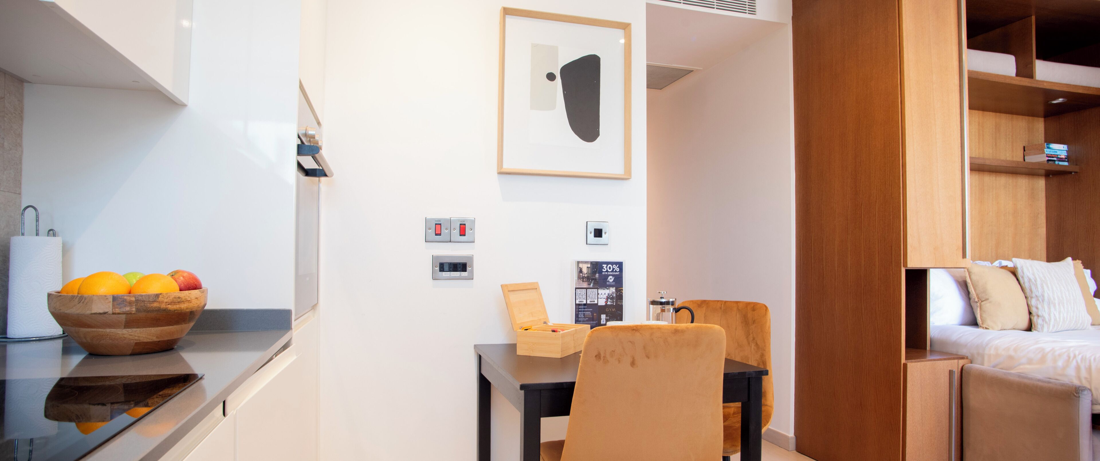 A modern hotel room with a wooden dining table, a fruit bowl, a small wooden side table, a bed with a wooden headboard, and a wall-mounted light switch.