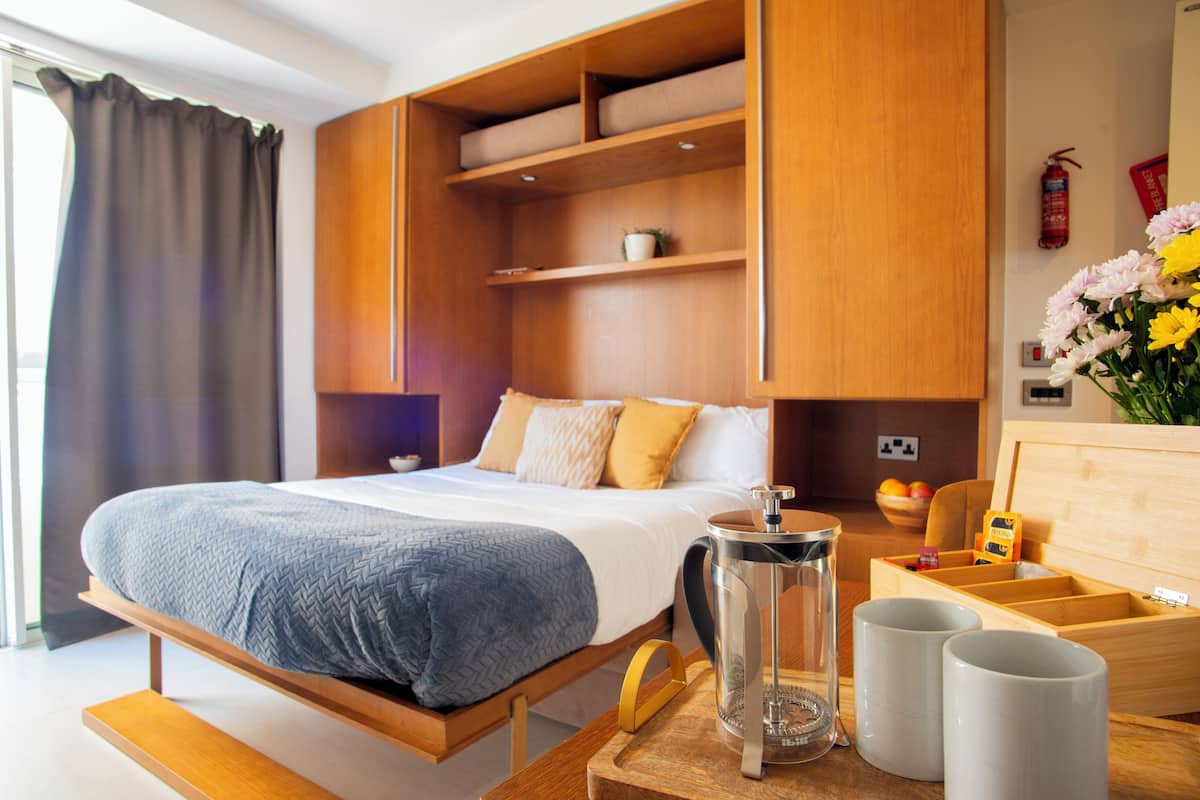 A modern hotel room with a bed, wooden shelving, a coffee maker, and a small table with cups.