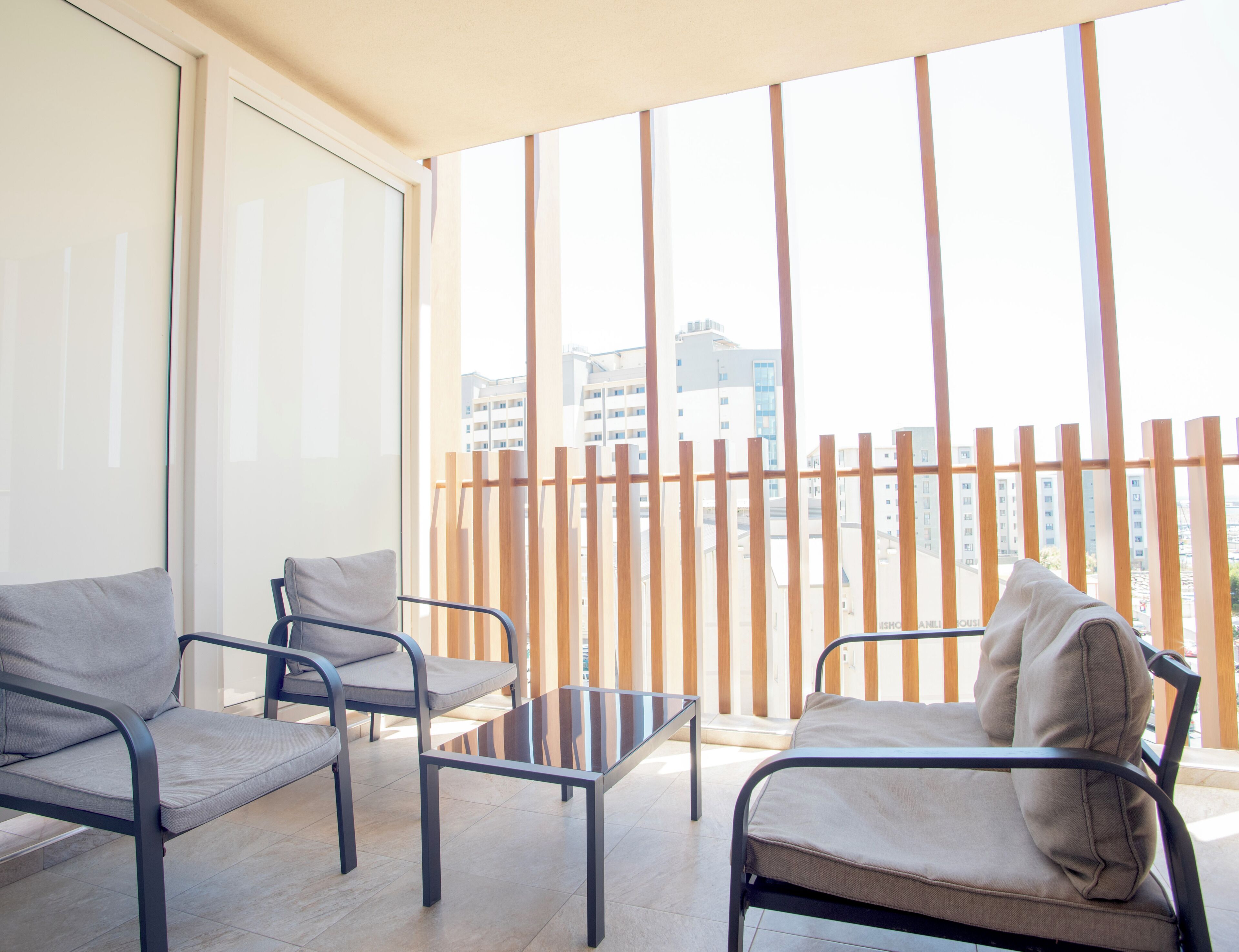 A balcony with two chairs, a small table, and a view of buildings through a wooden fence.