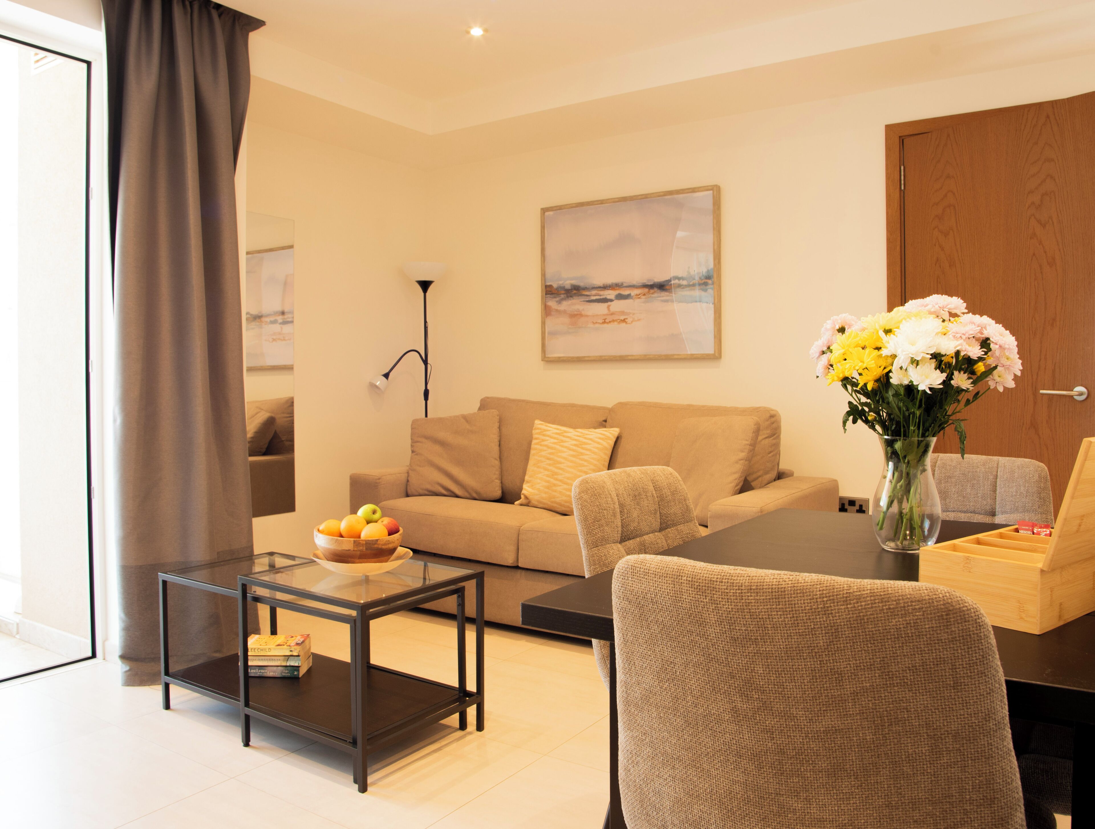 A modern living room with a sofa, a glass coffee table, a chair, a vase with flowers, and a painting on the wall.