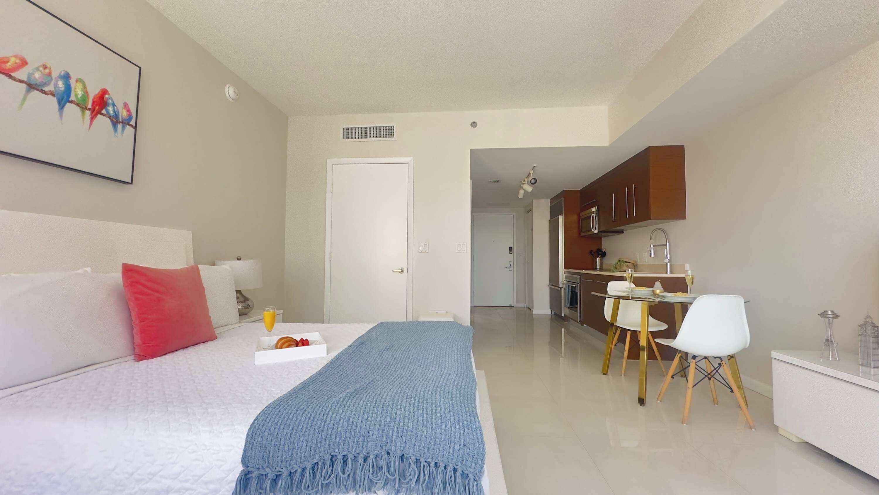 A hotel room with a bed, a bedside table with a glass of orange juice, a breakfast tray, a blue throw blanket, and a painting of birds on the wall.