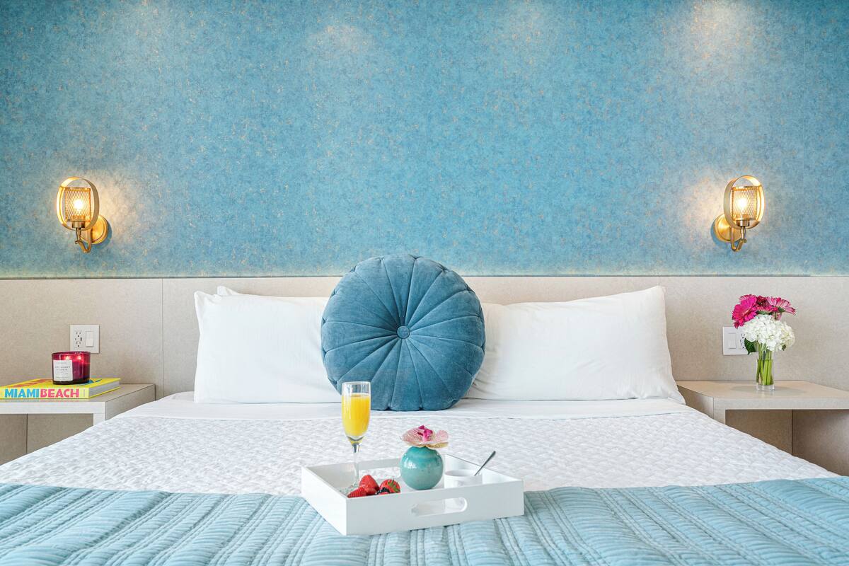A neatly made bed with a blue pillow, a bedside table with a book, a candle, and a tray with fruit and a glass of orange juice.