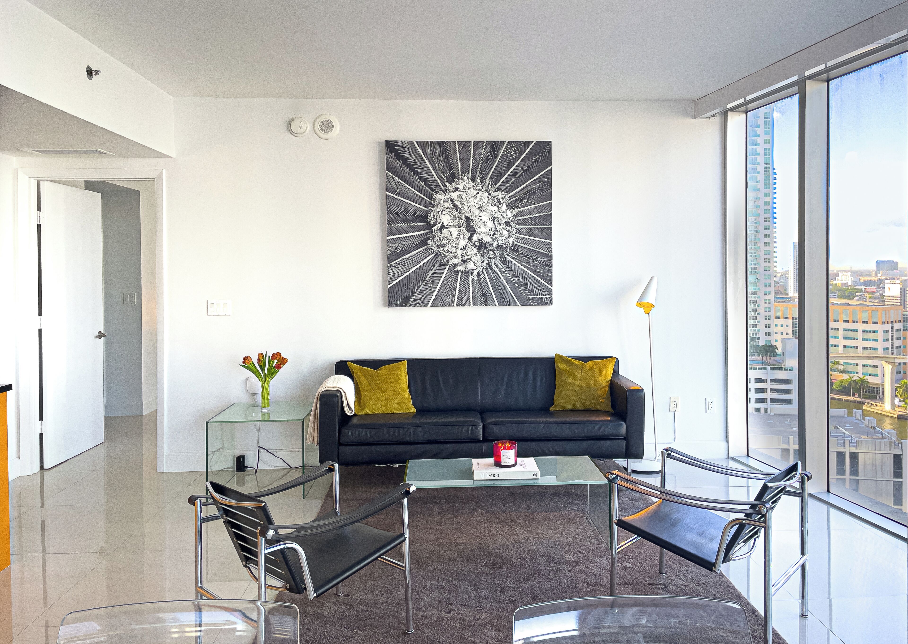 A modern living room with a black sofa, glass coffee table, and metal chairs. A large window offers a city view.