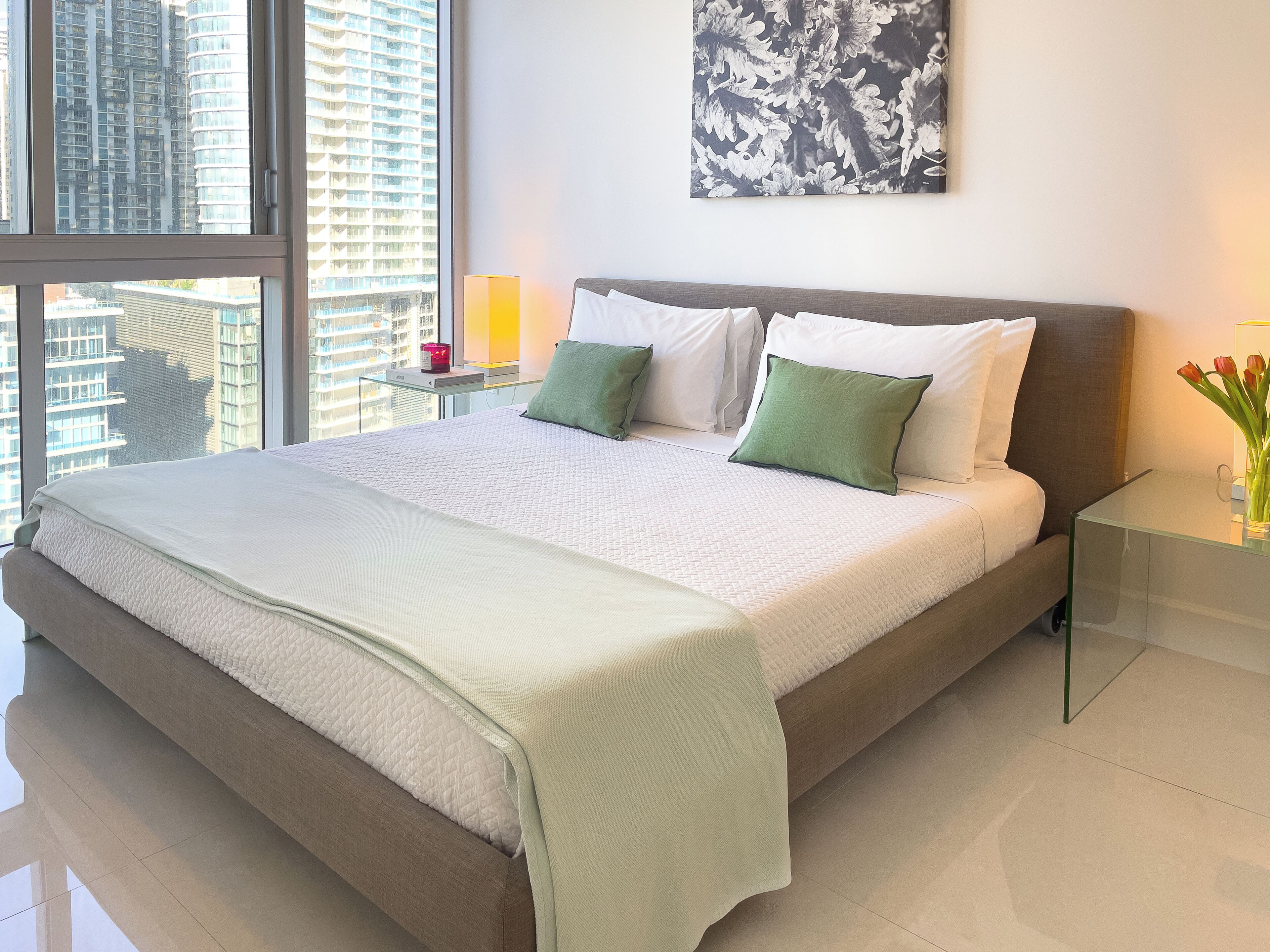 Quarto de hotel moderno com uma cama grande, uma mesa de cabeceira com abajur, vista da cidade através de grandes janelas e uma obra de arte na parede.