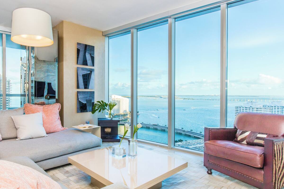 A modern living room with a sofa, armchair, and coffee table, large windows with a view of the water and buildings.