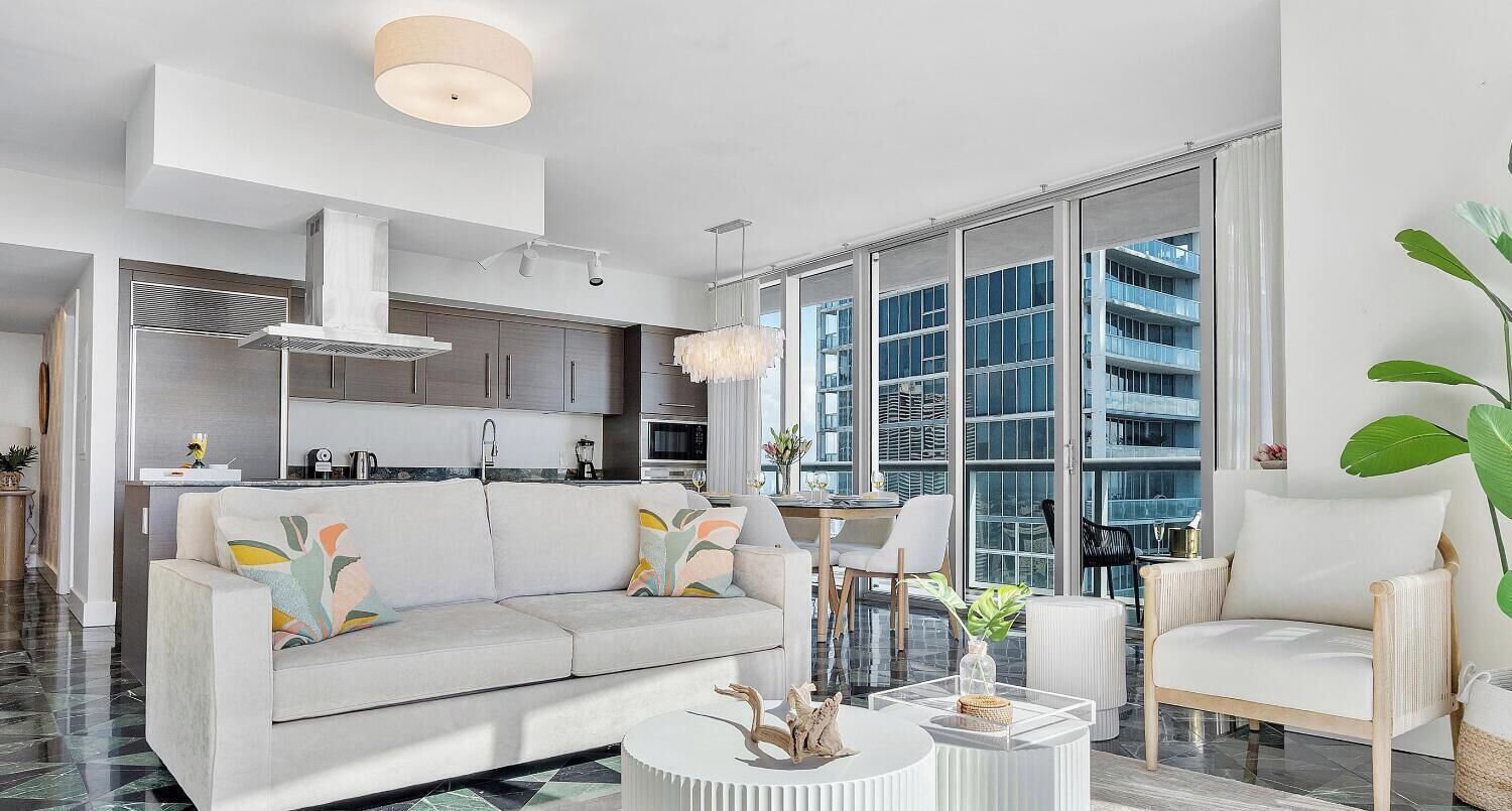 A modern living room with a sofa, coffee table, and chairs, a kitchen in the background, and large windows offering a view of the city.