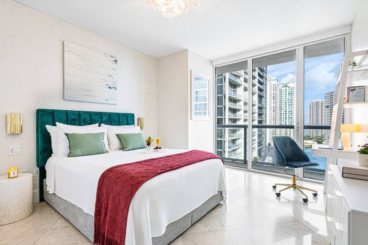 A modern hotel room with a large bed, a desk, a chair, and a view of high-rise buildings.