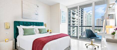 A modern hotel room with a large bed, a desk, a chair, and a view of high-rise buildings.