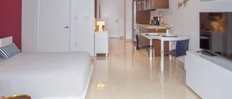 A modern hotel room with a bed, a small desk, a kitchenette, and a TV.