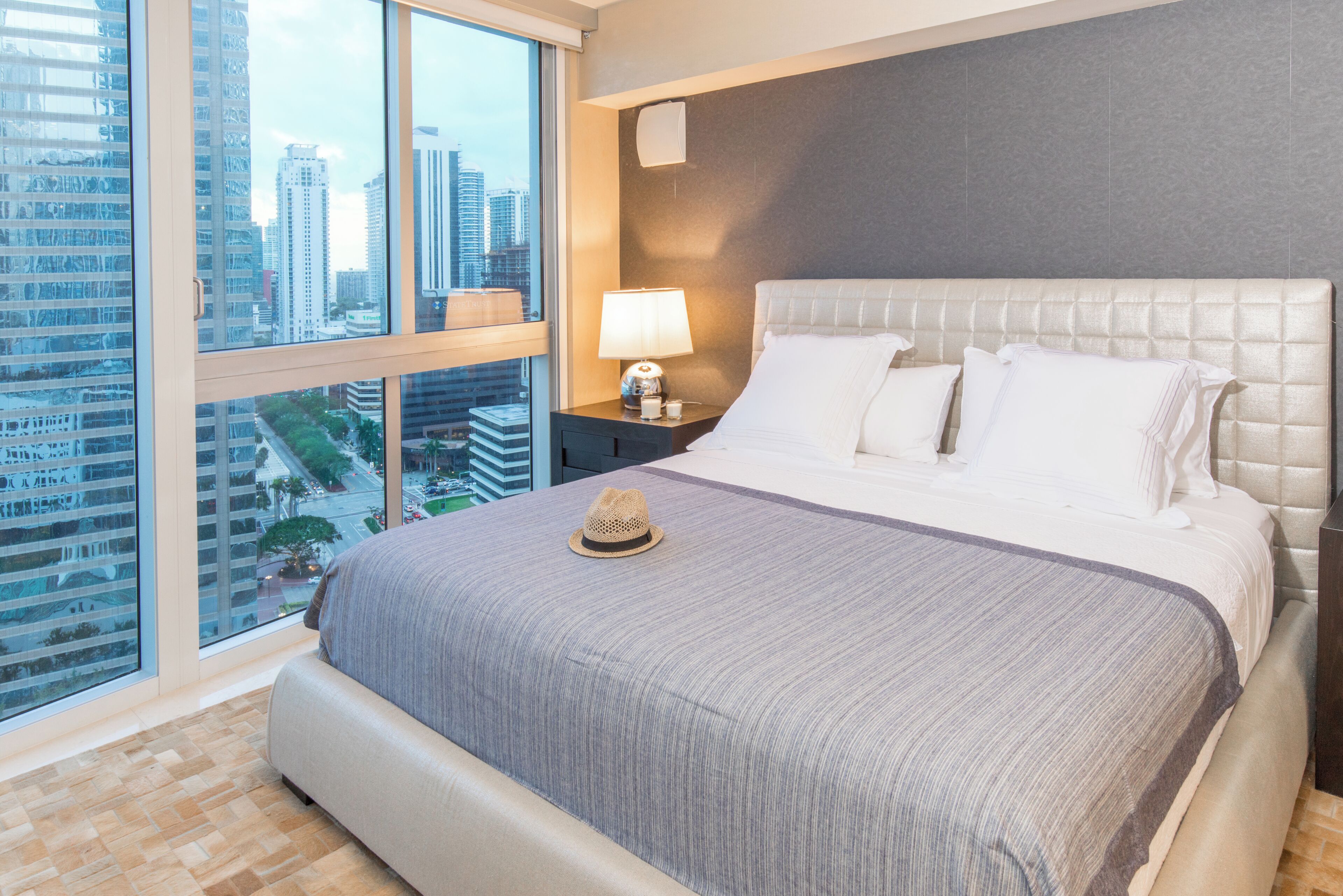 Habitación de hotel con un ventanal grande, una cama con una manta gris, una mesita de noche con una lámpara y vistas a la ciudad.
