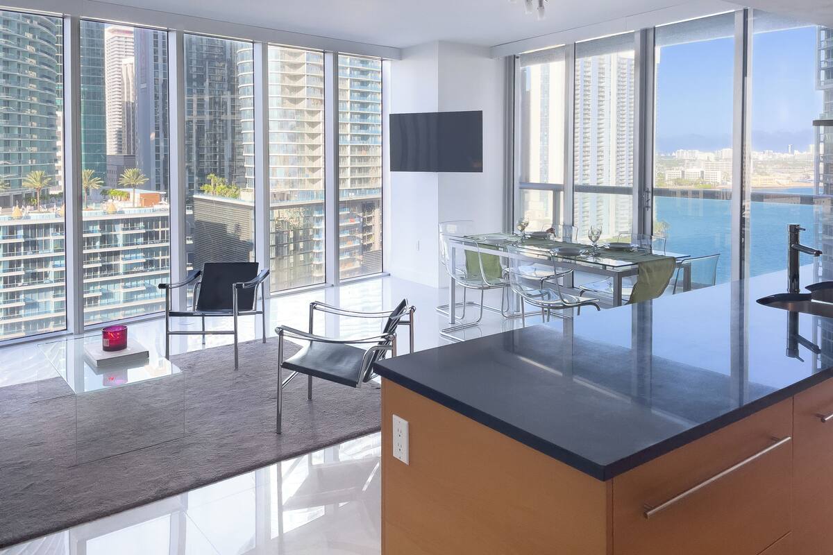 A modern kitchen with a glass island, dining area, and large windows offering city views.