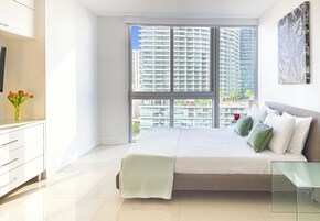 Un dormitorio moderno con un ventanal grande, una cama con almohadas, una mesita de noche y un televisor en la pared.