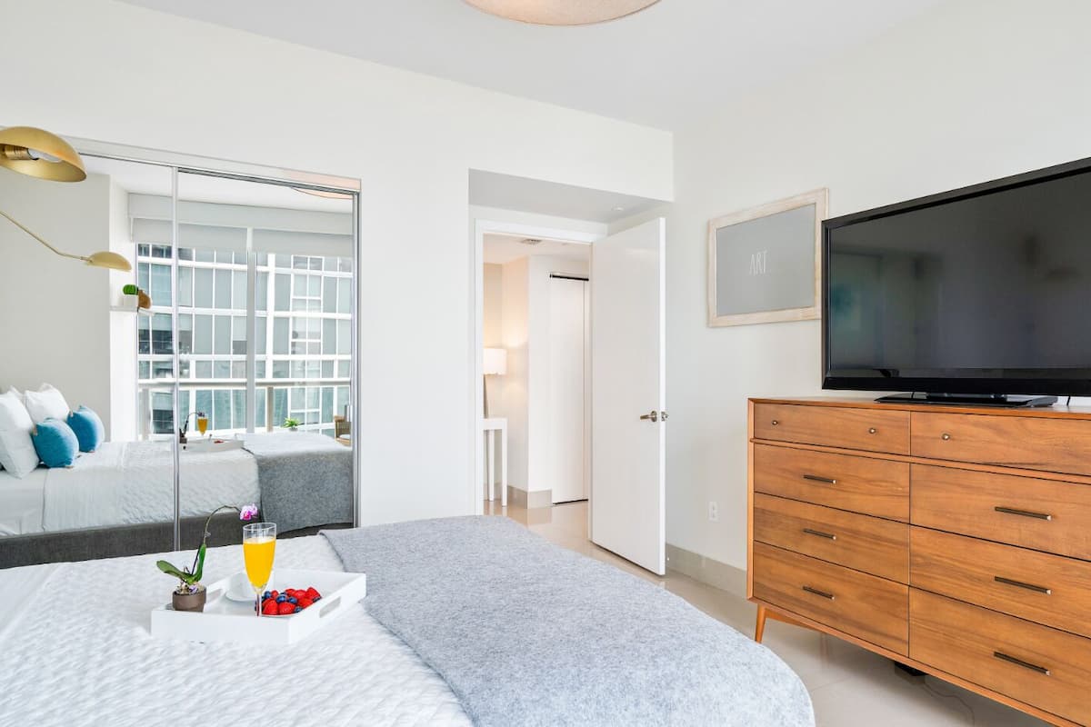 A modern bedroom with a bed, bedside table, TV, and a view of a balcony.