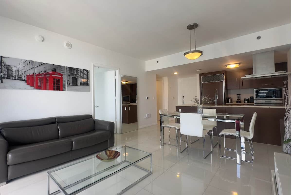 A modern living room with a sofa, glass coffee table, dining area with chairs, and a kitchen with cabinets and appliances.