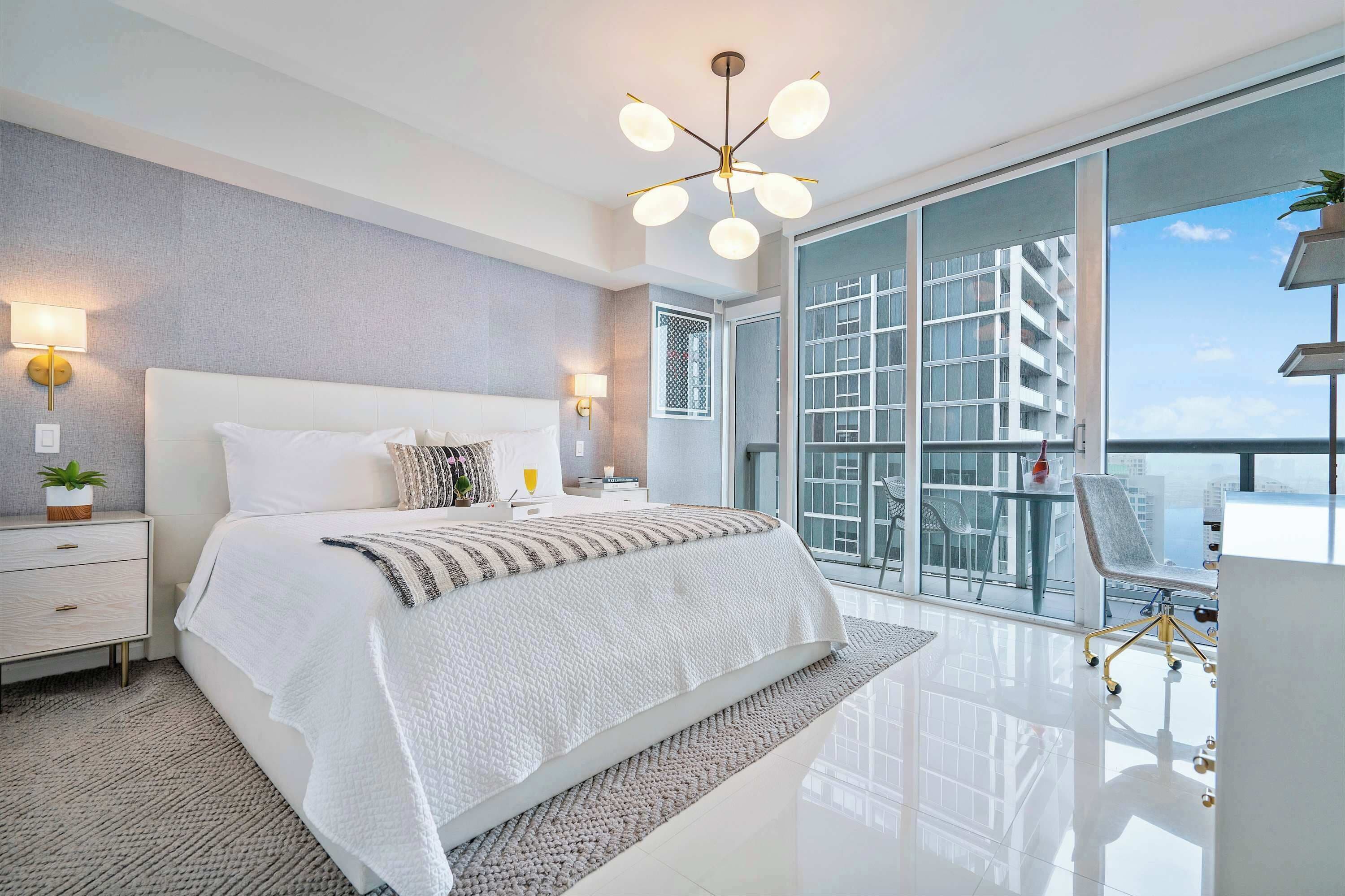 A modern bedroom with a large bed, bedside lamps, a view of a building through a glass door, and a balcony with a chair.