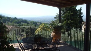 A covered outdoor patio with a round table and chairs, a large potted plant, and a view of a cityscape and mountains in the distance. - Camere Ramaccia (Assisi)
