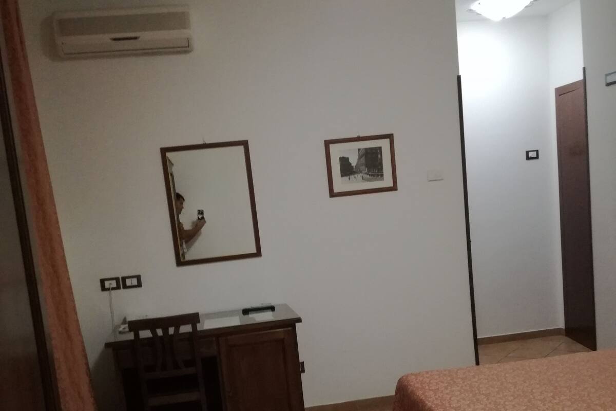 A room with a bed, a desk, and a chair. There is a mirror on the wall and a picture frame.