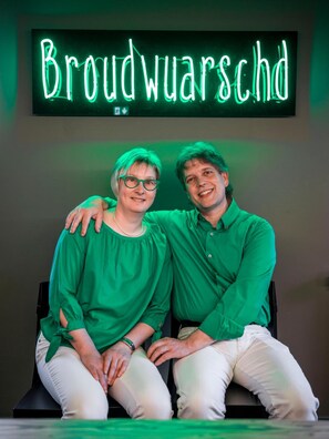 Zwei Personen sitzen vor einem leuchtenden Schild mit der Aufschrift 'Broudwarschd'.