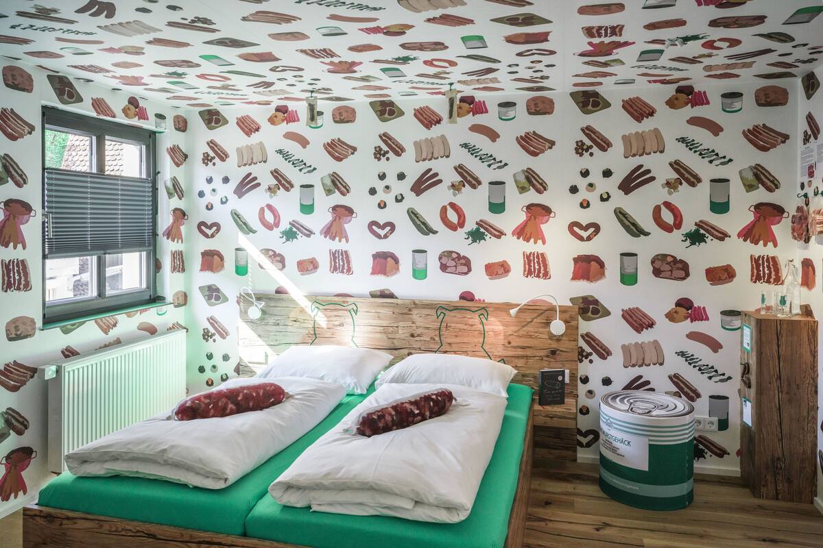 Ein Zimmer mit einem hölzernen Kopfteil, zwei Betten mit grüner Bettwäsche, einem Heizkörper, einem Fenster mit Jalousien und Wänden, die mit Illustrationen von Lebensmitteln verziert sind.