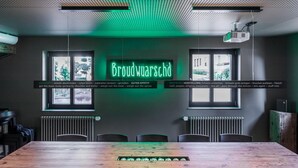 Une salle de conférence avec une longue table en bois, six chaises et un panneau lumineux au néon sur lequel on peut lire « Brodwurschd ».