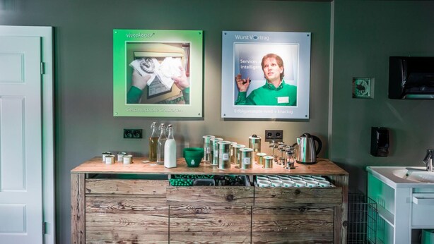 Un espace d’accueil avec un comptoir en bois, une machine à café et un évier. Des affiches encadrées ornent le mur, et une porte se trouve sur la gauche.