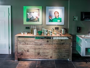 Ein Empfangsbereich mit einem Holzboden, einer Kaffeemaschine und einem Waschbecken. An der Wand hängen gerahmte Plakate und links ist eine Tür.