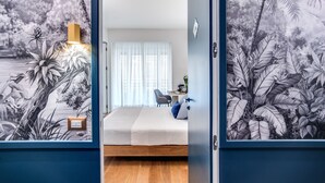 A bedroom with a bed, a desk, and a chair, a window with curtains, and a wall with a tropical mural. - Victor Boutique Hotel (Milan)