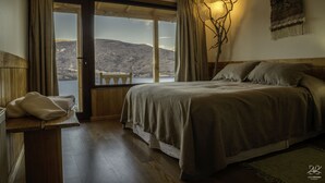 Een slaapkamer met een groot bed, een houten hoofdbord, een bankje bij het raam en uitzicht op een bergachtig landschap.