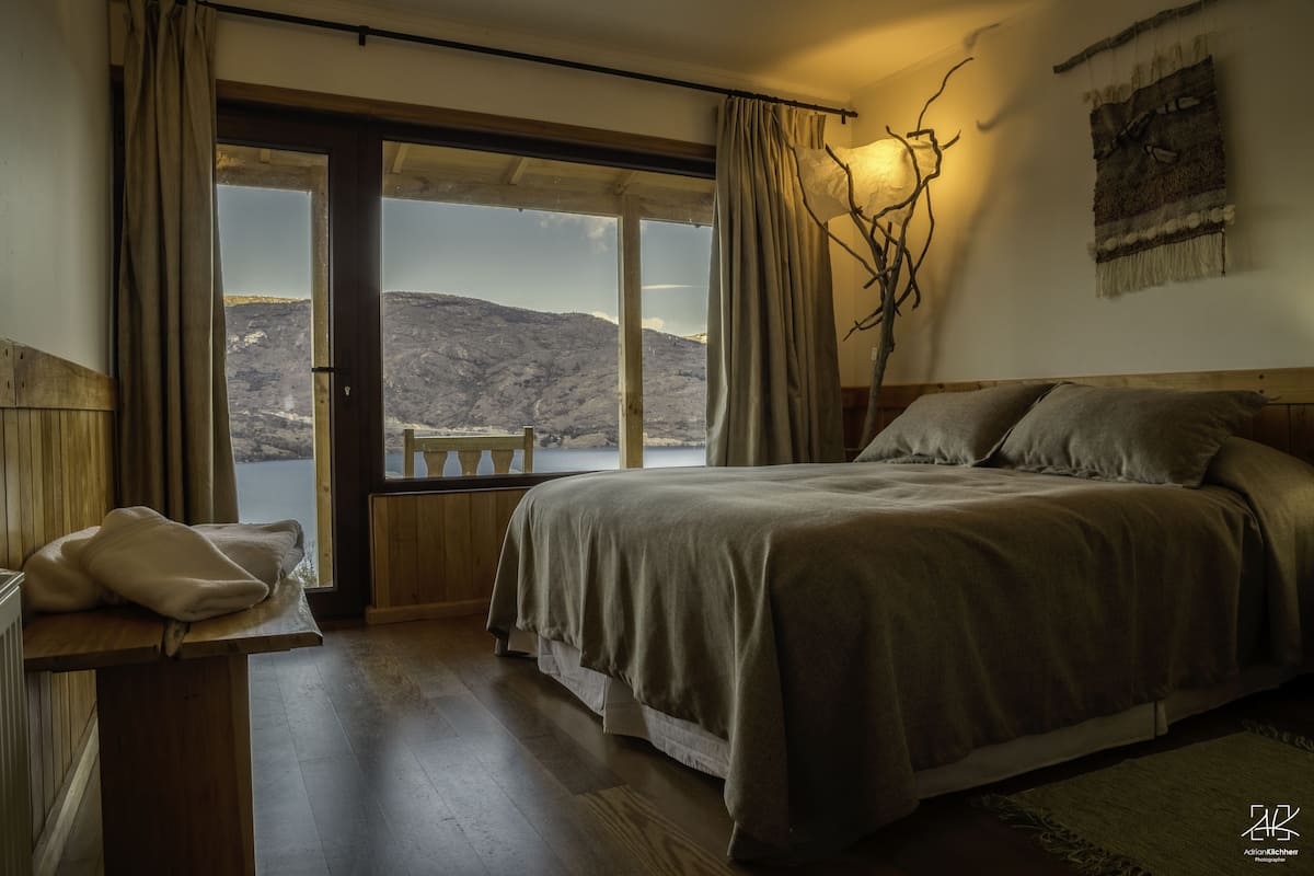 Une chambre à coucher avec un grand lit, une tête de lit en bois, un banc près de la fenêtre et une vue sur un paysage montagneux.