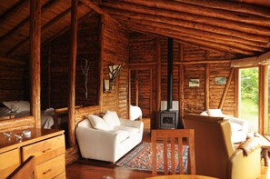 Interior de uma cabana de tronco com um sofá, cadeiras, uma mesa e uma lareira a lenha.
