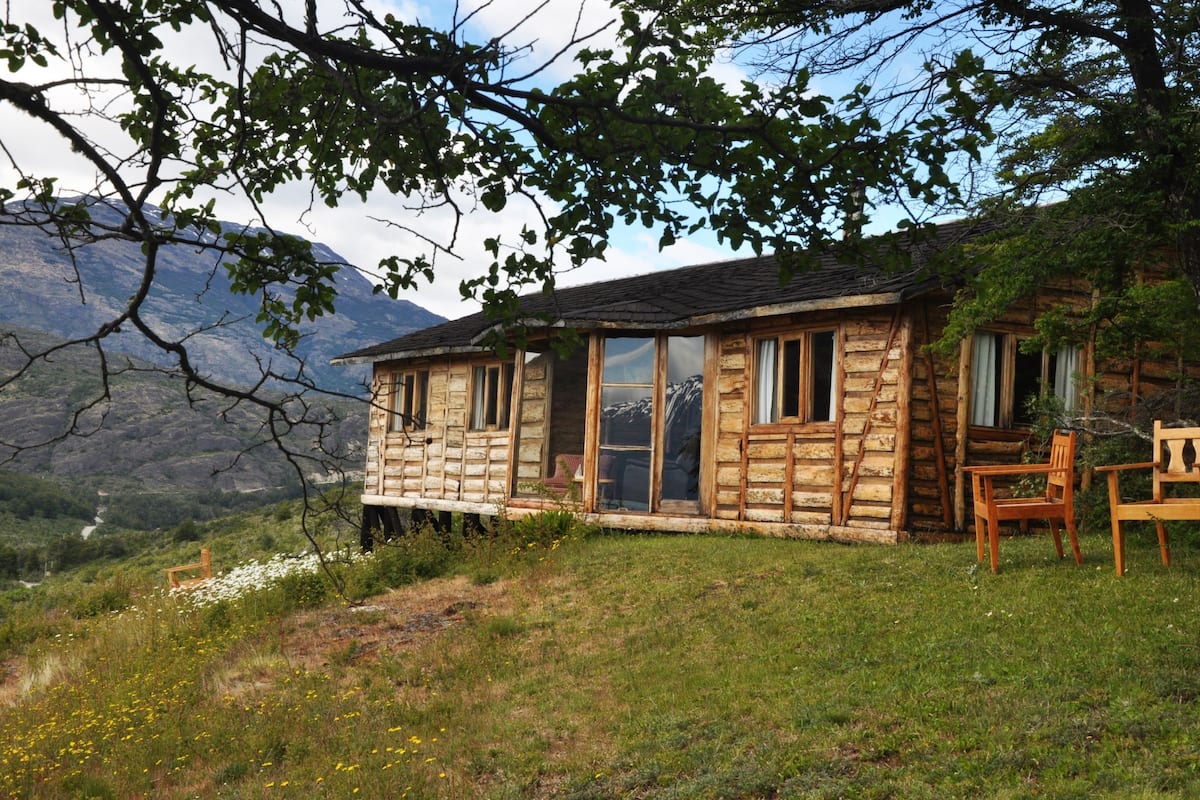 Un chalet rustique en rondins de bois, doté d’un porche en bois, de grandes fenêtres et offrant une vue sur un paysage montagneux.