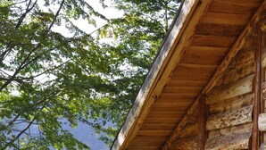 Een rustieke houten blokhut met een houten veranda en één raam, met uitzicht op een grasachtig landschap en een waterpartij.