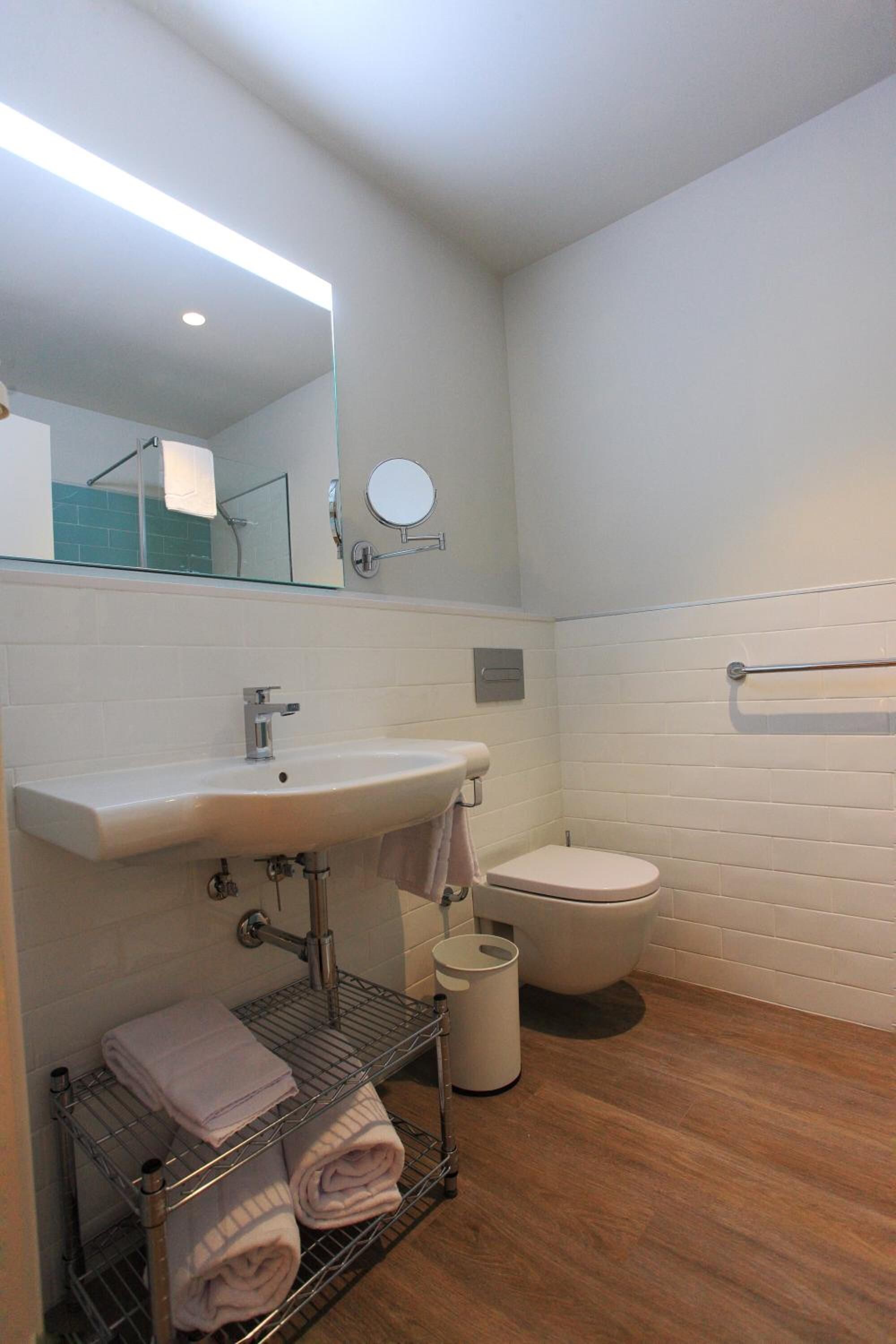 A bathroom with a white sink, mirror, and toilet. There is a towel rack and a metal shelf with towels.