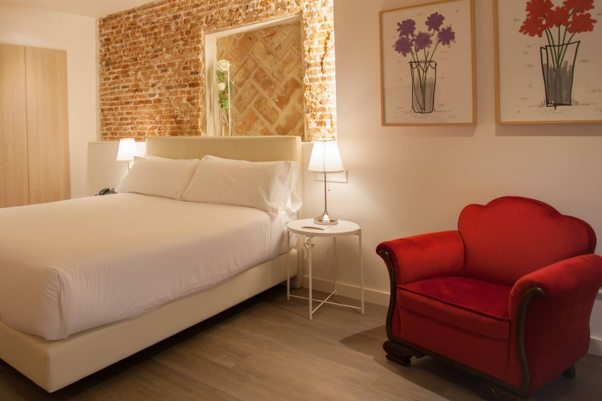 A hotel room with a large bed, a red armchair, a small table with a lamp, and artwork on the walls.