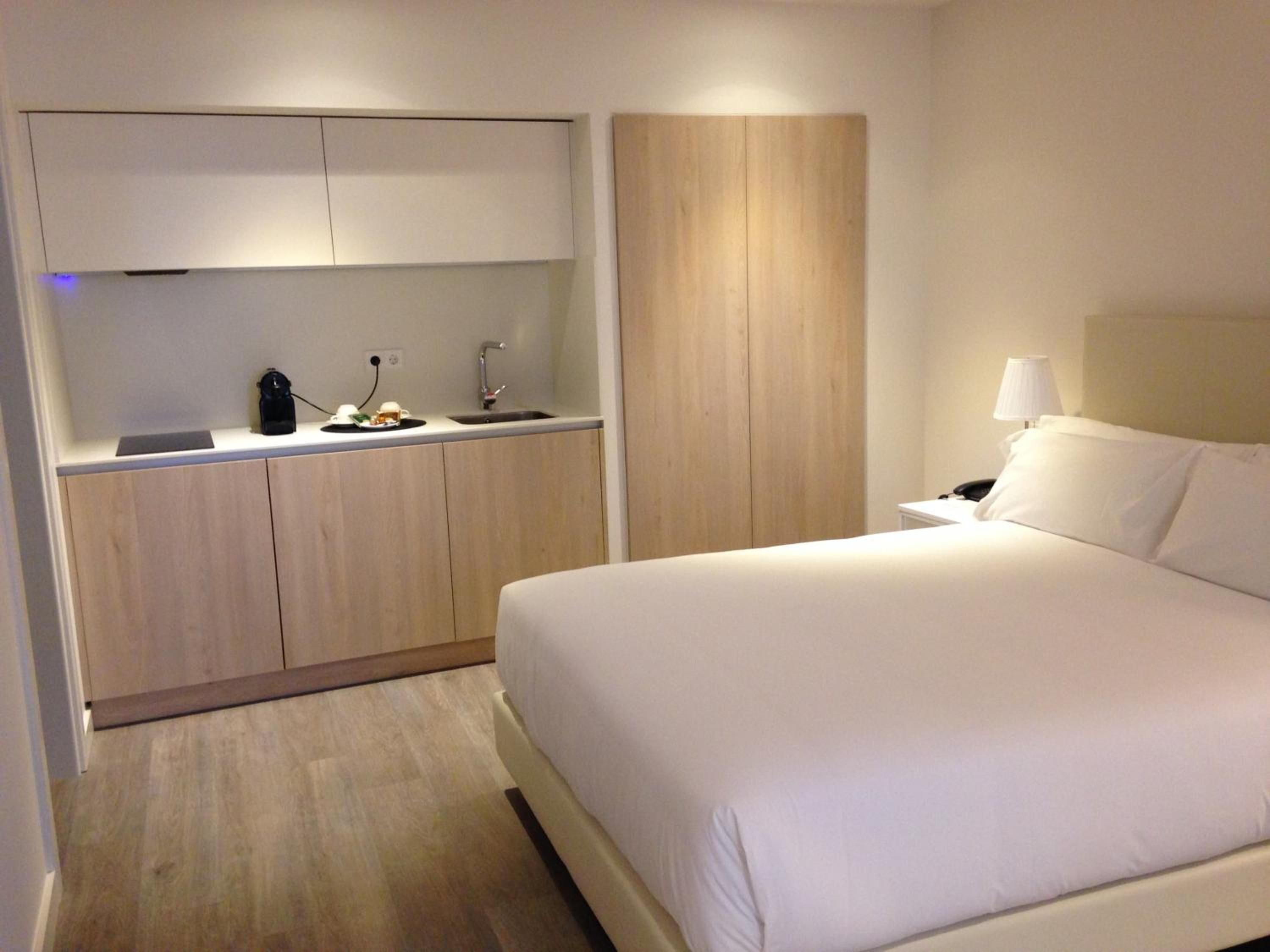 Una habitación de hotel pequeña con cama, kitchenette y mesita de noche con lámpara.