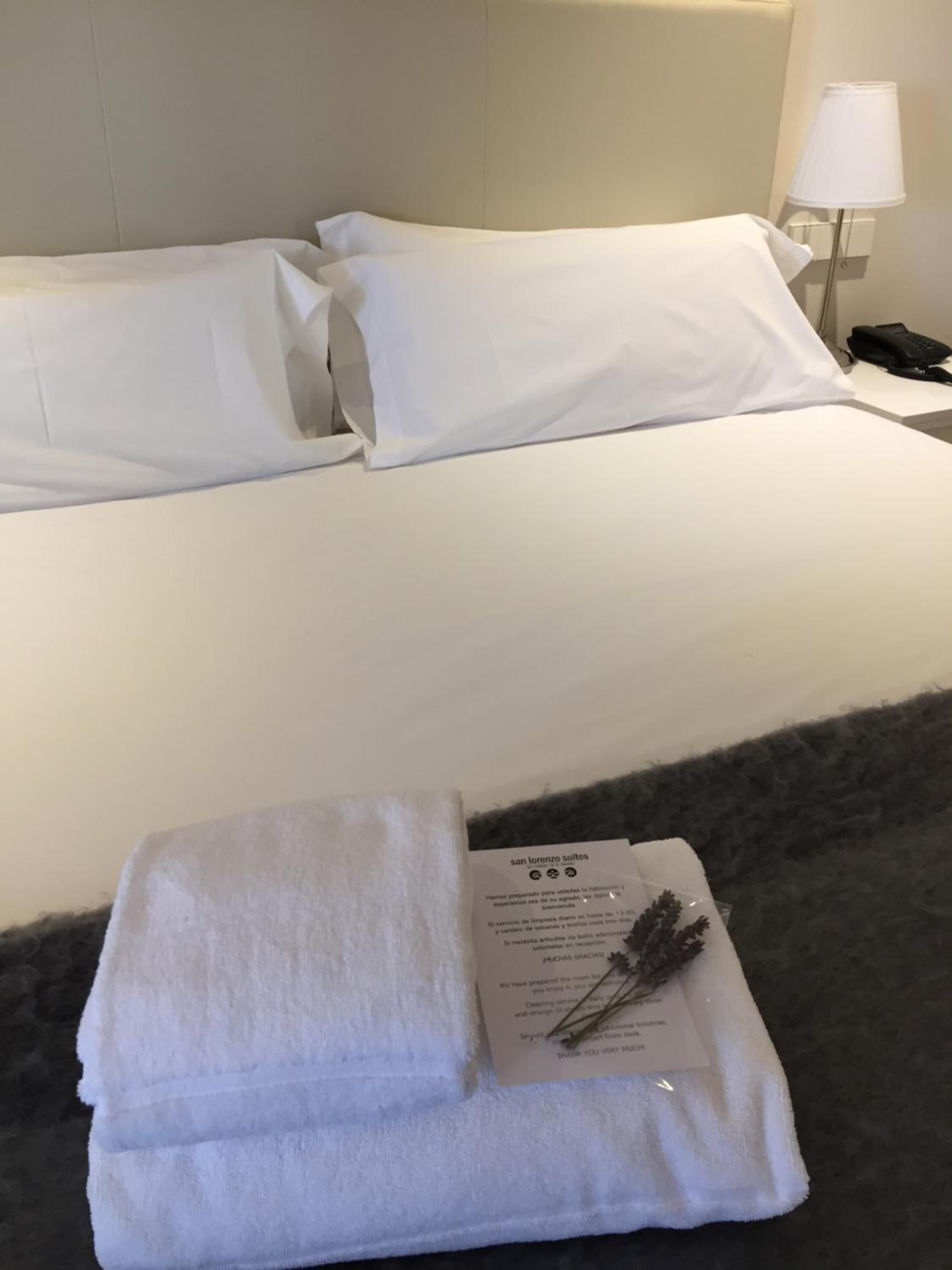Una cama bien tendida con sábanas y almohadas blancas, una toalla blanca doblada y una tarjeta con texto sobre un suelo alfombrado oscuro.