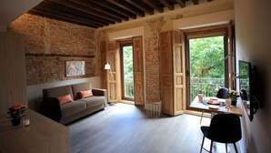 A cozy living room with a sofa, a dining table, and a TV. There is a window with a view of greenery outside. - San Lorenzo Suites (San Lorenzo de El Escorial)