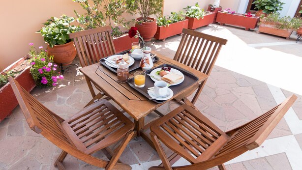 Una mesa de madera dispuesta con alimentos para el desayuno, rodeada de sillas de madera, en un patio al aire libre con plantas en macetas.
