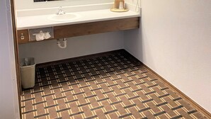 Un baño con lavamanos blanco, espejo y secador de pelo. El suelo tiene un diseño y hay un bote de basura pequeño.