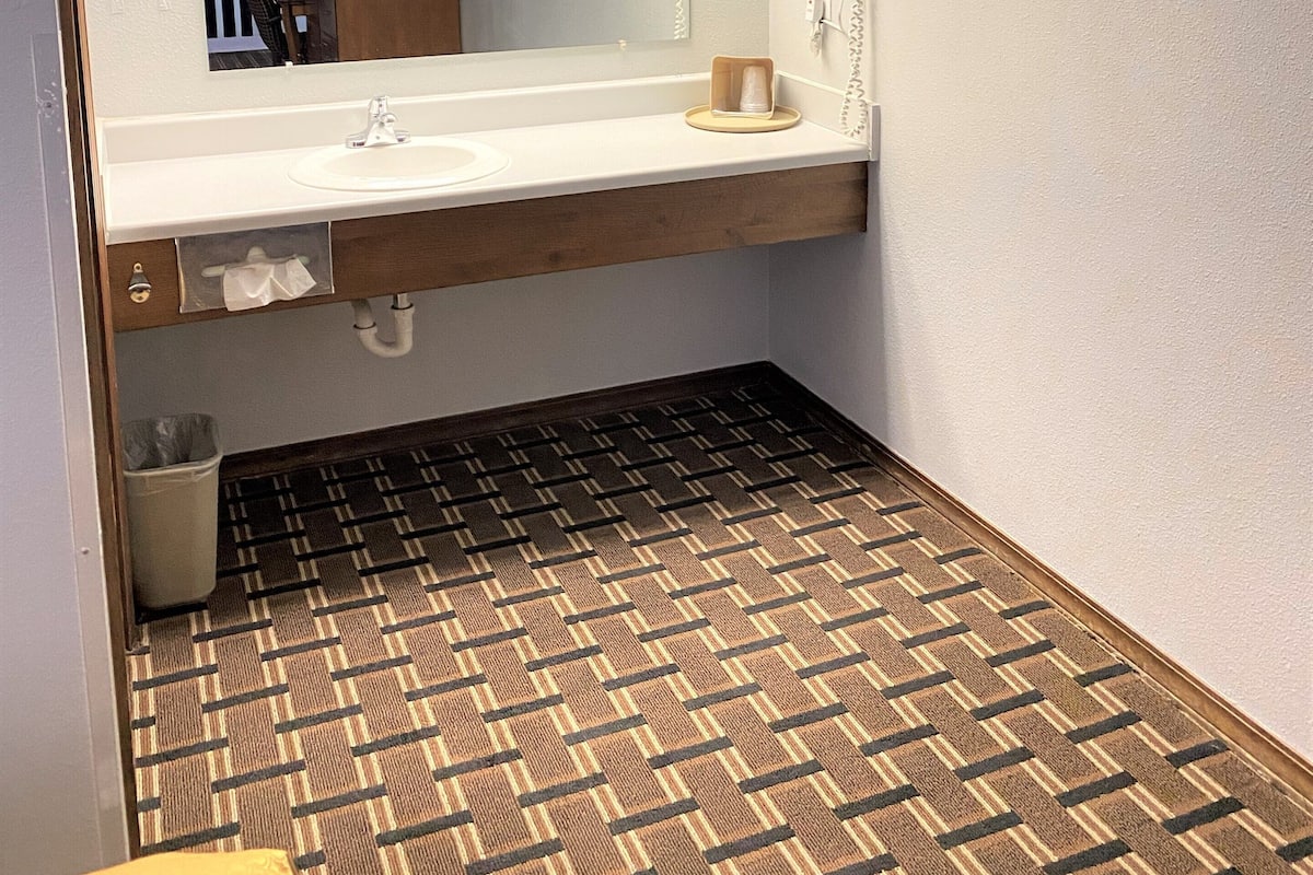 Un baño con lavamanos blanco, espejo y secador de pelo. El suelo tiene un diseño y hay un bote de basura pequeño.