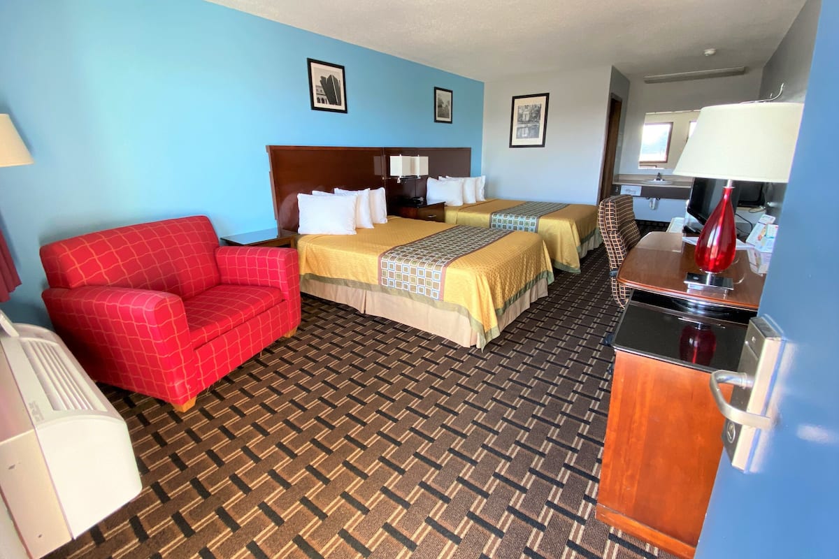 Una habitación de hotel con dos camas, un sillón rojo, un escritorio con lámpara y una alfombra estampada.