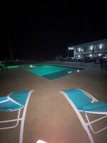 Zona de piscina por la noche con dos sillones reclinables y un edificio al fondo.