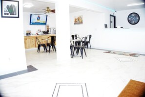 A modern kitchen with wooden cabinets, a dining area with black chairs, a ceiling fan, and a wall clock.