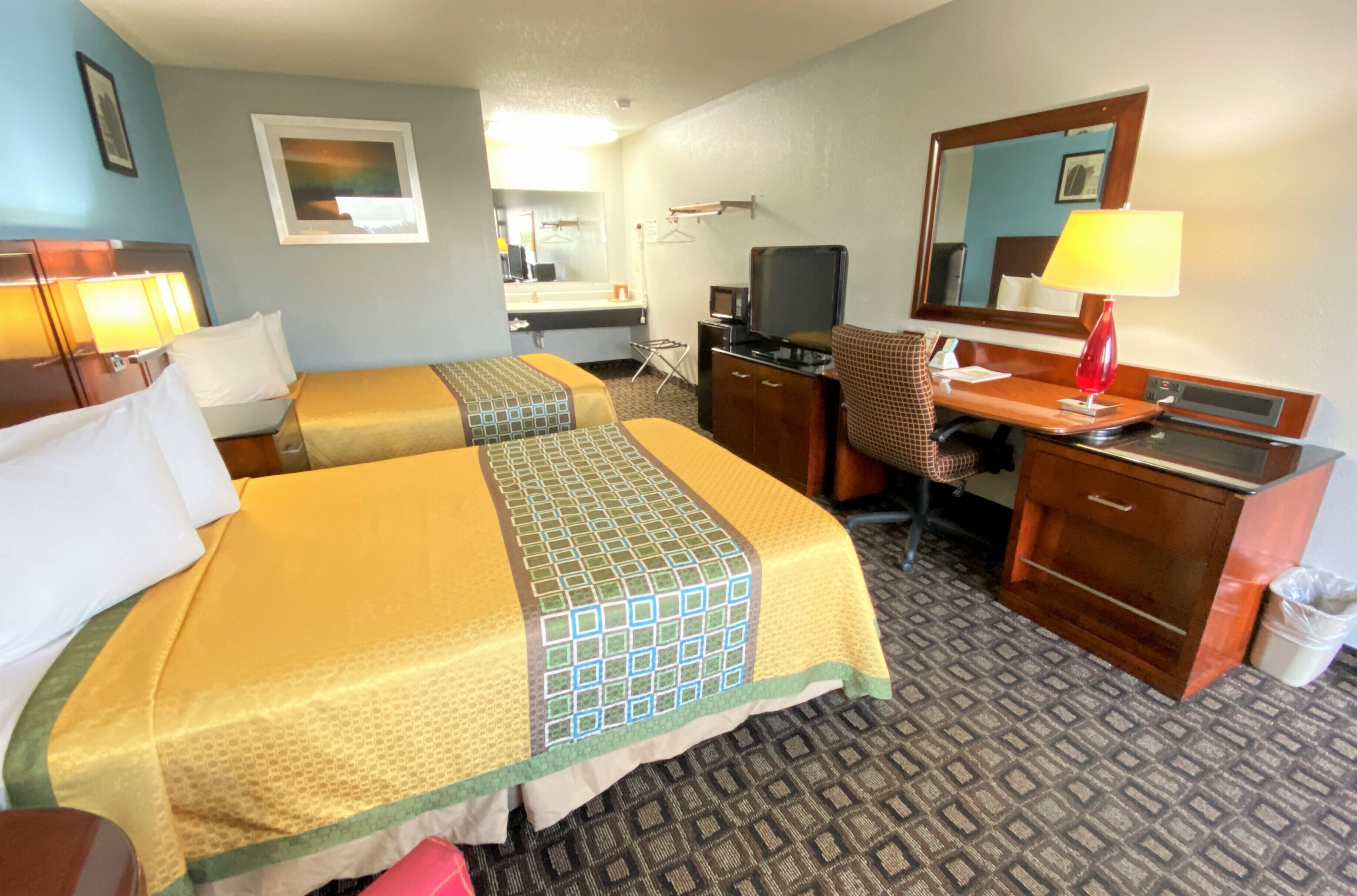 A hotel room with two beds, a desk, a chair, a TV, a mirror, and a lamp.