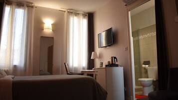 Habitación de hotel con cama, escritorio, silla, televisor y baño visible a través de una puerta abierta.