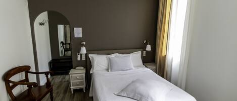 Una habitación de hotel con una cama, una silla de madera, una mesita de noche, una lámpara y un espejo.